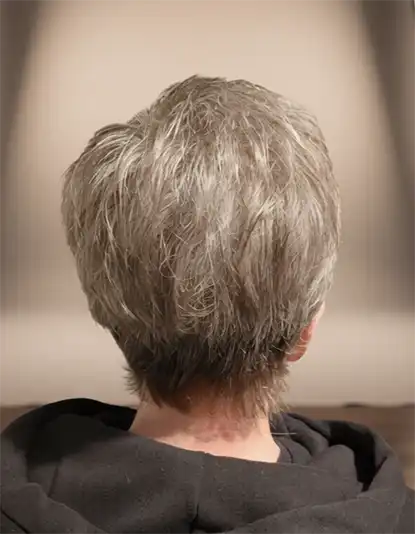 Back view of a person with short, grey hair wearing a black hoodie, facing a neutral, softly lit background.