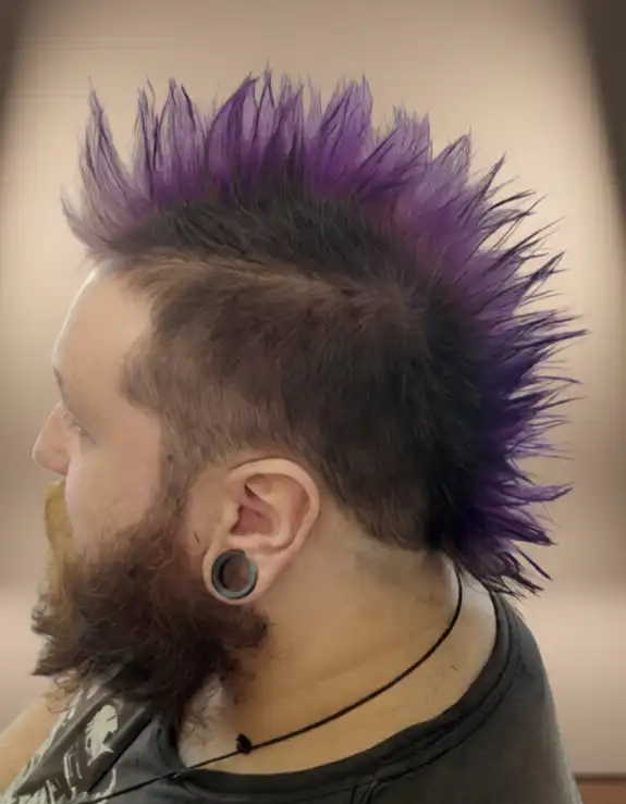 A person with a purple spiked mohican hairstyle, dark beard, stretched earlobe, and wearing a sleeveless shirt and necklace.