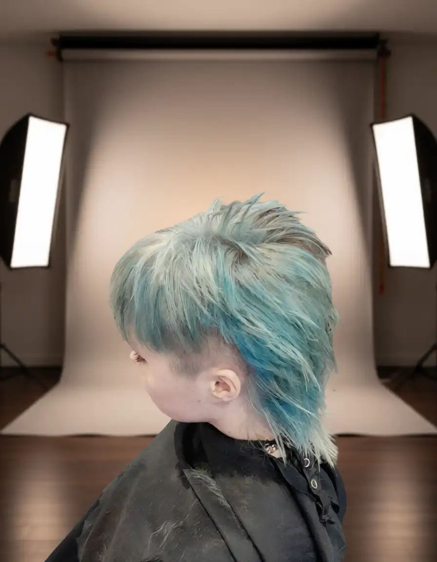 Person with short, spiky, blue mullet hairstyle posing in a photography studio with softbox lights and a neutral backdrop.