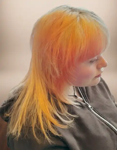 Person with layered hair dyed in orange and yellow shades, wearing a grey zip-up hoodie, looking downward.