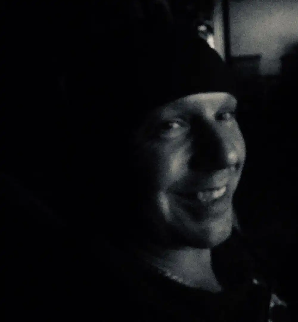 Iain Laidlaw wearing a beanie smiles in a dimly lit, black-and-white photo.