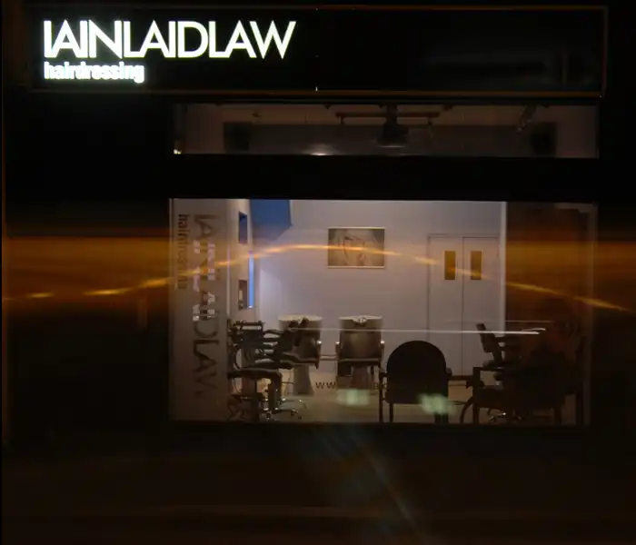A dimly lit hair salon named IAINLAIDLAW hairdressing with empty chairs visible through the front window at night.